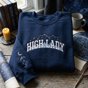 May include: Navy blue sweatshirt with the embroidered text "HIGH LADY" in white, with a constellation design above. The left sleeve features a moon, mountains, and stars. A cozy, celestial-themed garment.
