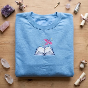 Puede incluir: Sudadera azul claro con un dise&ntilde;o bordado de un libro abierto, un drag&oacute;n rosa y estrellas. La sudadera est&aacute; hecha de un material suave y es perfecta para uso casual. El dise&ntilde;o es detallado y colorido.