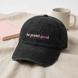 May include: Black baseball cap featuring the embroidered text "be pretty good" in yellow, pink, and purple. The cap has a curved peak and a button on top. A pink notebook and a white mug are visible in the background.