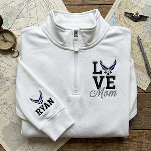 May include: White quarter-zip sweatshirt with "LOVE Mom" embroidered in black and grey. The sweatshirt features a blue and gold Air Force emblem. The sleeve has the name "RYAN" embroidered in black with an Air Force emblem.