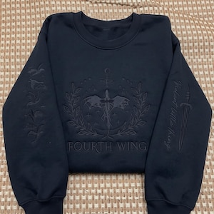 May include: A black crewneck sweatshirt featuring tonal embroidery. The chest displays a dragon within a laurel wreath, stars, and the text "FOURTH WING". The left sleeve shows birds, while the right sleeve features a dagger and "Violent Little Thing".