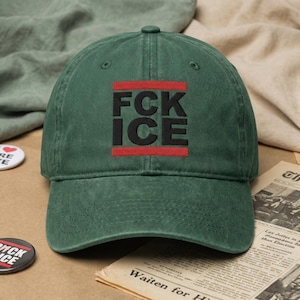 May include: A dark green baseball cap featuring the text "FCK ICE" in black, with red lines above and below. The cap is made of a soft, textured fabric and has a curved peak. The hat is on a brown surface.