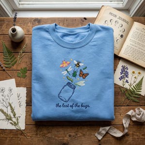 May include: A light blue crewneck sweatshirt featuring an embroidered design of a glass jar with various colourful insects like a butterfly, moth, bee, and dragonfly floating above it, with the text "the last of the bugs" stitched below.