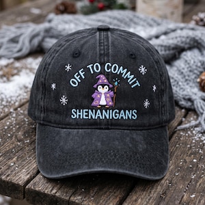 May include: A charcoal gray baseball cap featuring an embroidered design of a penguin dressed as a wizard in a purple robe and hat, holding a staff. The text reads OFF TO COMMIT SHENANIGANS in light blue, surrounded by white embroidered snowflakes.