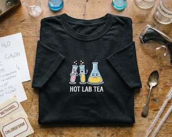 May include: A black short-sleeve t-shirt featuring a graphic of two smiling test tubes and one grumpy Erlenmeyer flask filled with colored liquids. Below the illustration, the text reads "HOT LAB TEA" in white capital letters.