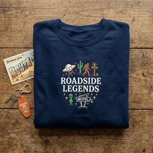 May include: A navy blue crewneck sweatshirt featuring an embroidered design with the text "ROADSIDE LEGENDS" and "MOTEL". The graphic includes a UFO, a cactus, a Bigfoot figure, a wooden signpost, a green alien, and small stars.