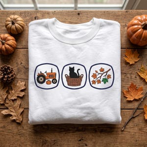 May include: A white crewneck sweatshirt featuring three distinct circular emblems with dark blue borders. The first emblem shows an orange tractor and a pumpkin, the second a black cat in a brown basket, and the third a branch with autumn leaves.