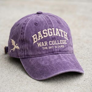 May include: A purple baseball cap with the text "BASGIATH WAR COLLEGE THE SKY IS OURS" embroidered in off-white. A dragon graphic is on the side. The cap has a curved brim and an adjustable strap.