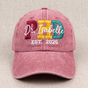 May include: A dusty rose baseball cap featuring embroidered text. The letters PHD are stitched in maroon, yellow, and teal. The white cursive text Dr. Isabelle is layered over the letters, with the white text EST. 2026 stitched below the main design.