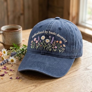 May include: A navy blue baseball cap with the embroidered phrase "radicalized by basic decency" above a floral design. The flowers include daisies, lavender, and other colourful blooms. The hat is on a wooden surface.