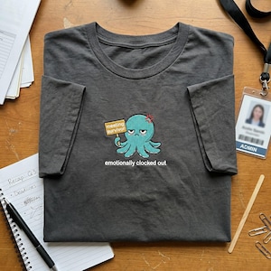 May include: Dark gray t-shirt featuring an embroidered teal octopus holding a "meeting survivor" sign, with the text "emotionally clocked out" below. The octopus has an angry expression. The shirt is folded on a wooden surface.
