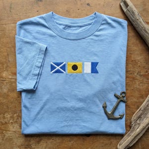 May include: A light blue short-sleeve t-shirt featuring an embroidered design of three nautical signal flags in the center. The flags are blue and white with a diagonal cross, yellow with a black circle, and white and blue with a swallowtail.