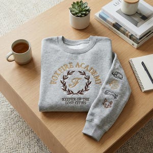 May include: A gray sweatshirt with embroidered text and patches. The front features "FOXFIRE ACADEMY" in gold lettering with a laurel wreath and the text "KEEPER OF THE LOST CITIES." The sleeve has patches of an elephant, a lion, a cheetah, and a fox.