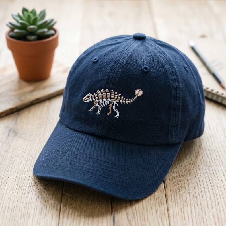 May include: A navy blue baseball cap featuring an embroidered design of an ankylosaurus dinosaur skeleton in shades of tan and white. The cap has a curved brim, a button on the top, and stitched eyelets for ventilation on the crown panels.