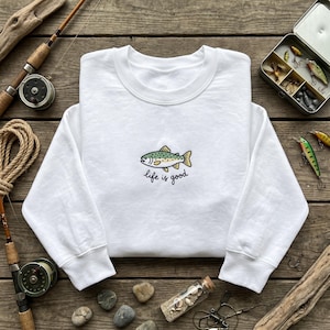 May include: White sweatshirt with a crew neck featuring an embroidered fish design in green, yellow, and blue, with the text "life is good" below. Fishing gear, including rods and lures, surrounds the sweatshirt on a wooden surface.