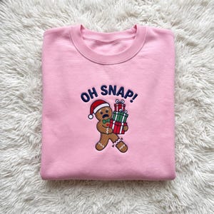 May include: A light pink crewneck sweatshirt featuring an embroidered design of a gingerbread person wearing a Santa hat and a green bow tie. The gingerbread person holds a stack of three wrapped gifts and has a broken leg. The text OH SNAP! is embroidered in dark blue above the figure.