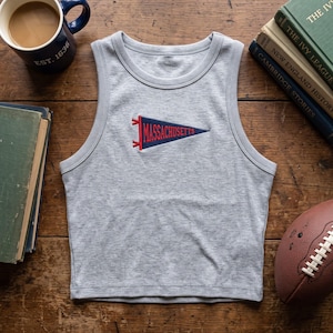 May include: A light grey ribbed tank top featuring a centered embroidered pennant graphic in navy blue and red. The pennant displays the text MASSACHUSETTS in red capital letters. The garment has a round neckline and sleeveless armholes.