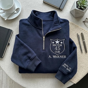 Peut inclure: Un sweat-shirt bleu marine à fermeture éclair quart de col avec un écusson brodé blanc indiquant "SECTOR 45 CCR" et "A. WARNER". La manche porte l'inscription "Ignite, my love, Ignite". Une tasse, des stylos et un carnet sont visibles.