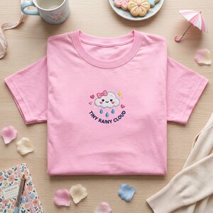 May include: A pink t-shirt with a round neck featuring an embroidered cloud design with a bow, hearts, and raindrops. The text "Tiny Rainy Cloud" is below the cloud. Other items include a teacup, biscuits, and a notebook.