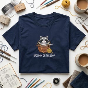 May include: Navy blue t-shirt featuring a cartoon raccoon crocheting in a basket, with the text "RACCOON ON THE LOOP" below. The image is surrounded by crochet hooks, yarn, and craft supplies.