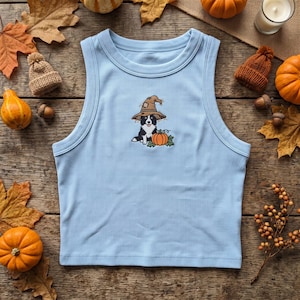 May include: A light blue ribbed tank top featuring an embroidered graphic of a black and white dog wearing a brown witch hat and sitting next to an orange pumpkin. The garment has a crew neckline and a sleeveless design with ribbed fabric.