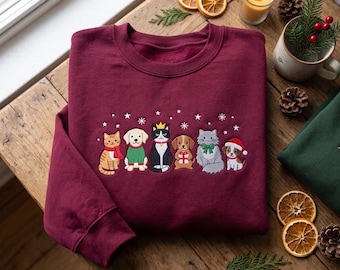 May include: A maroon crewneck sweatshirt featuring an embroidered graphic of six pets, including cats and dogs, wearing holiday accessories like scarves, sweaters, and a Santa hat, surrounded by small white embroidered snowflakes.