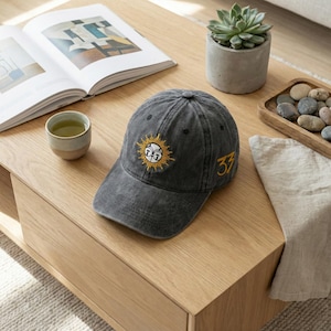 May include: A charcoal grey baseball cap with a sun and face design embroidered in yellow. The number 33 is embroidered in yellow on the side. The hat is on a light wood table.