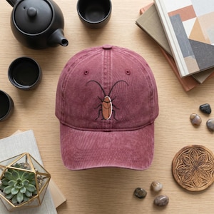 May include: A maroon baseball cap featuring an embroidered graphic of a brown cockroach with long black antennae centred on the front panel. The cap has a structured crown, a curved brim, and visible stitching details across the fabric surface.