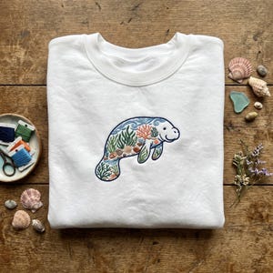 May include: A white crewneck sweatshirt featuring a central embroidered design of a manatee. The manatee's body is filled with a detailed pattern of colorful coral, seaweed, and various seashells in shades of orange, green, and brown.