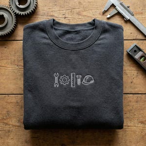 May include: Charcoal grey crew neck sweatshirt featuring white embroidered engineering icons: a spanner, cogwheel, ruler, bolt, and hard hat. The sweatshirt is folded on a wooden surface.