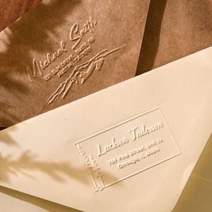 Puede incluir: Primer plano de dos sobres con texto en relieve. Uno es marrón claro con el nombre "Michael Smith" y la dirección. El otro es blanquecino con el nombre "Jackson Anderson" y la dirección. Los sobres están sobre una superficie de madera.