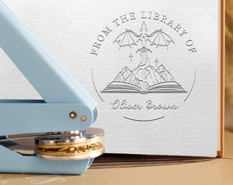 May include: A light blue metal embosser tool positioned next to a white paper page featuring a circular embossed design. The design includes a sword, dragon wings, mountains, an open book, and the text "FROM THE LIBRARY OF Oliver Brown."