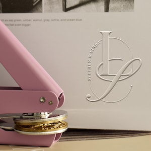 May include: A pink embossing seal with a gold embossed design. The seal is pressing down on a gold embossed design. The text "STEPHEN & LINDA" is on the right side of the image.