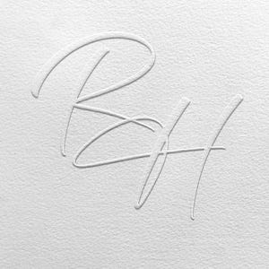Może przedstawiać: Białe, wytłoczone inicjały „RH” na tle teksturowanego papieru. Litery są w delikatnym, kursywnym kroju pisma, tworząc subtelny, elegancki design. Ogólna estetyka jest minimalistyczna i czysta, z naciskiem na teksturę i formę.