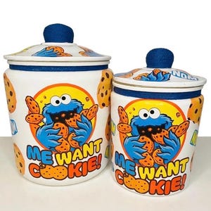 Op de afbeelding: Twee witte keramische voorraadpotten met deksel, voorzien van het blauwe Koekiemonster dat chocolate chip cookies eet. De tekst ME WANT COOKIE! staat in blauw en geel gedrukt, samen met NOM-tekst en koekjespatronen op de buitenkant.