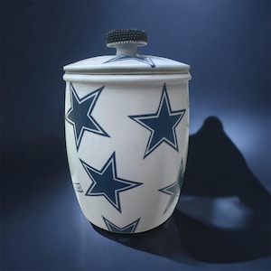 Könnte beinhalten: Weiße Keramikdose mit einem blauen Sternenmuster und einem schwarzen Strassdeckel. Die Dose hat eine abgerundete Form und eine glatte Oberfläche.