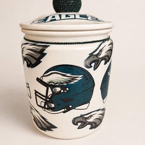 May include: A white ceramic jar with a lid, decorated with Philadelphia Eagles football team graphics. The jar features a teal football helmet and eagle head designs. The lid has the word "EAGLES" and a beaded knob.