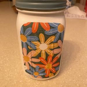 Op de afbeelding: Witte weckpot met een teal deksel. De pot heeft een kleurrijk bloemmotief met oranje, gele en blauwe bloemen op een donkergroene achtergrond. Het wordt waarschijnlijk gebruikt voor opslag of decoratie.