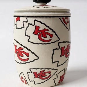 May include: A cream-colored ceramic jar with a lid, adorned with red and black Kansas City Chiefs arrowhead logos. The lid features a black jeweled knob, and the rim has a black beaded trim. A decorative storage container.
