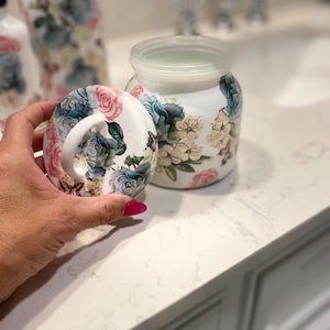 Peut inclure: Pot en céramique blanc avec couvercle transparent et motif floral avec des fleurs bleues, roses et crème. Le couvercle du pot est retiré, révélant l'intérieur. Un couvercle assorti avec une poignée est tenu à proximité, également décoré de fleurs.