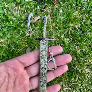 Può includere: Un portachiavi in metallo a forma di spada con un intricato motivo in rilievo sul fodero e sull'impugnatura. Il pomolo ha la forma di una testa di lupo e una piccola gemma rosso brillante è incastonata al centro della guardia. Un anello è incluso.