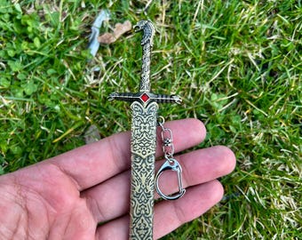 Puede incluir: Un llavero de metal con forma de espada que presenta un intrincado patrón en relieve en la vaina y la empuñadura. El pomo tiene forma de cabeza de lobo y una pequeña piedra preciosa de color rojo brillante está engastada en el centro de la guarda.