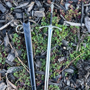 Puede incluir: Una espada plateada con una empu&ntilde;adura en forma de cruz y un mango negro, junto a una vaina negra con un anillo. La espada est&aacute; sobre un lecho de astillas de madera y plantas verdes.