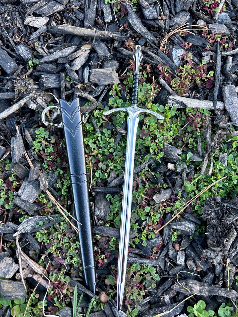 Puede incluir: Una espada plateada con una empu&ntilde;adura en forma de cruz y un mango negro, junto a una vaina negra con un anillo. La espada est&aacute; sobre un lecho de astillas de madera y plantas verdes.