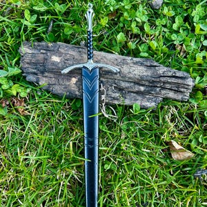 Puede incluir: Una espada decorativa con una vaina negra y detalles plateados, apoyada sobre un trozo de madera. La espada presenta una empu&ntilde;adura y una guarda ornamentadas. El fondo es hierba verde.