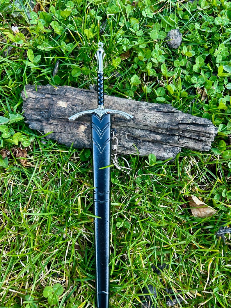 Puede incluir: Una espada decorativa con una vaina negra y detalles plateados, apoyada sobre un trozo de madera. La espada presenta una empu&ntilde;adura y una guarda ornamentadas. El fondo es hierba verde.