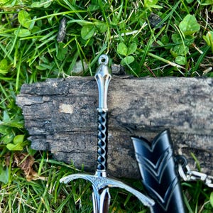 Puede incluir: Una espada plateada en miniatura con un mango negro retorcido y una vaina negra. La espada descansa sobre un trozo de madera desgastada, rodeada de hierba verde y tr&eacute;bol.