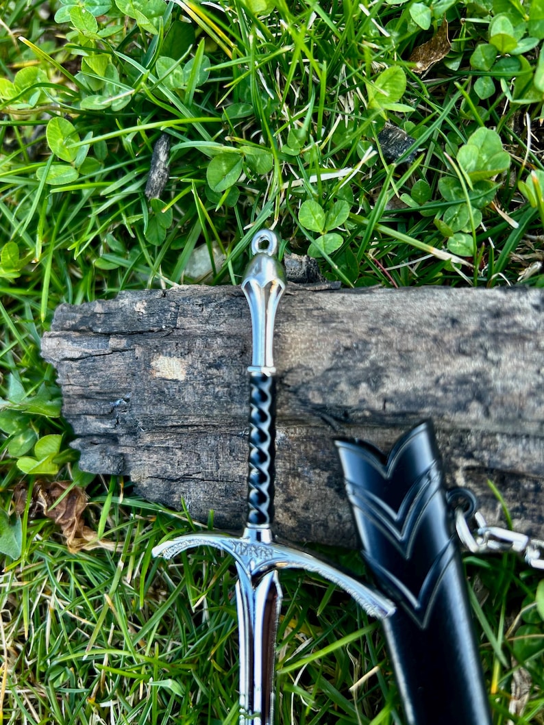 Puede incluir: Una espada plateada en miniatura con un mango negro retorcido y una vaina negra. La espada descansa sobre un trozo de madera desgastada, rodeada de hierba verde y tr&eacute;bol.