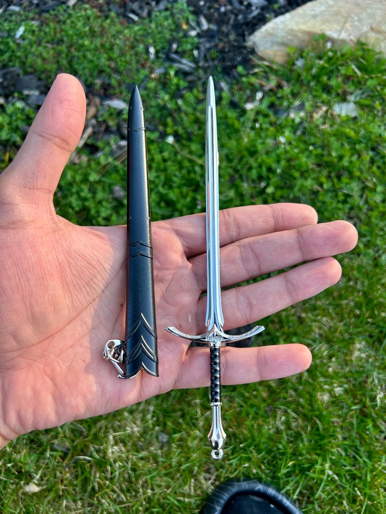 Puede incluir: Una espada decorativa en miniatura con una hoja plateada y un mango negro, junto a su vaina negra. La espada presenta detalles intrincados en la empu&ntilde;adura y el pomo. La vaina tiene un cierre plateado decorativo. Los art&iacute;culos se muestran sobre un fondo de hierba verde.