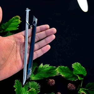 Puede incluir: Una espada plateada en miniatura con un mango y una hoja ornamentados, junto a una vaina negra. La espada se sostiene en una mano, con hojas verdes y bayas oscuras en el fondo. El dise&ntilde;o de la espada es detallado.